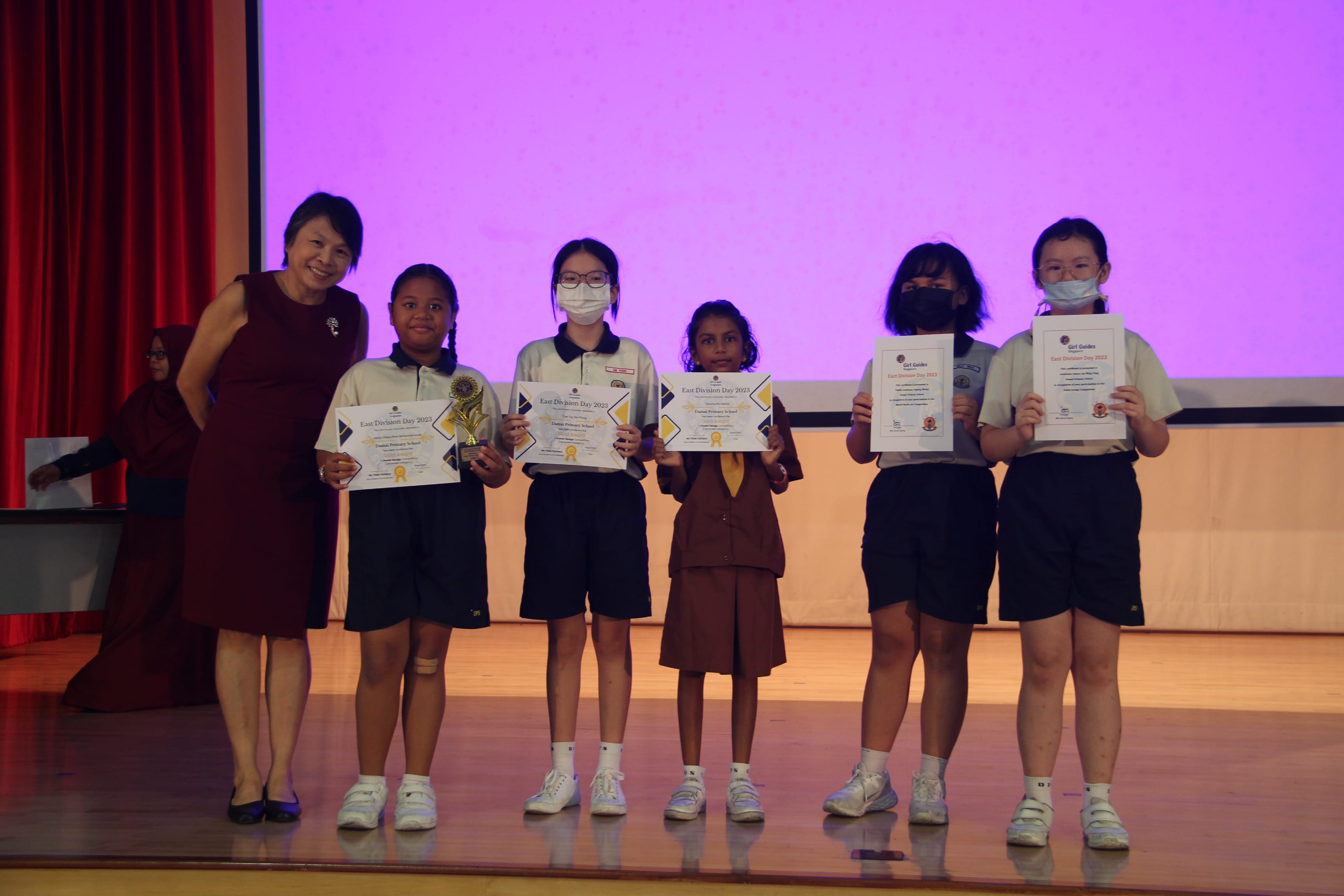 Five young students holding certificates stand on a stage with an adult woman at an East Division Day award ceremony.